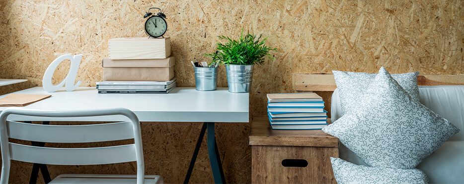 Brown chipboard-looking wall of a small, cluttered bedroom with a bed on the right and desk space on the left with a white metal chair and books, stationary on desk